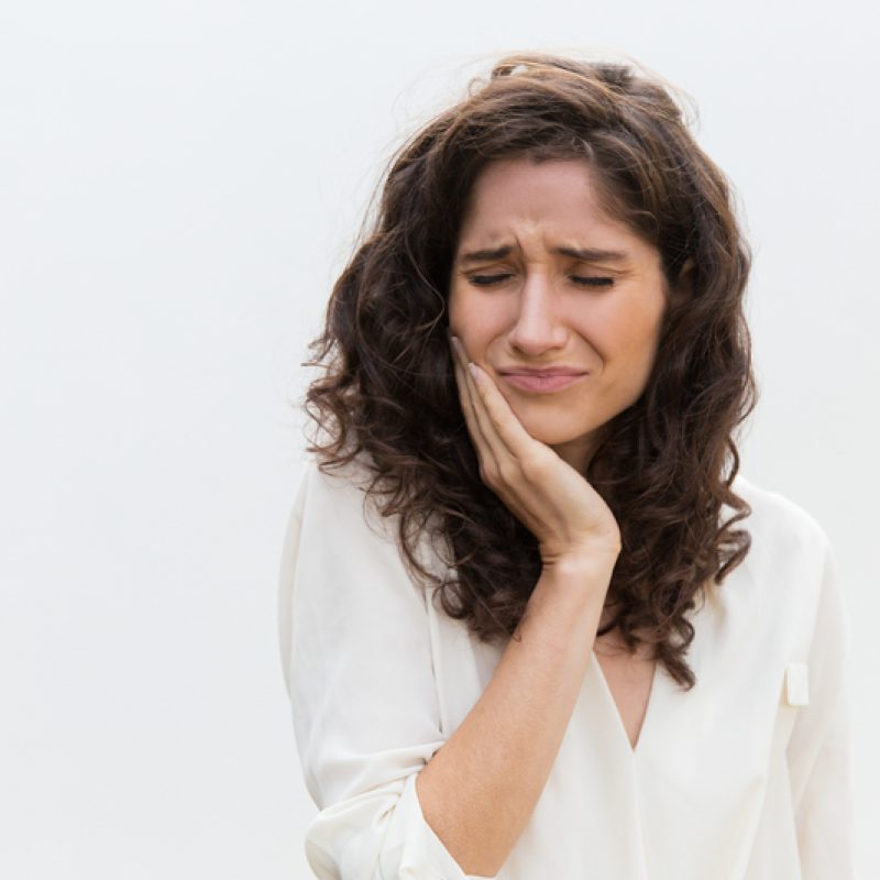 Frustrated unhappy woman suffering from toothache. Wavy haired young woman in casual shirt standing isolated over white background. Dental problem concept